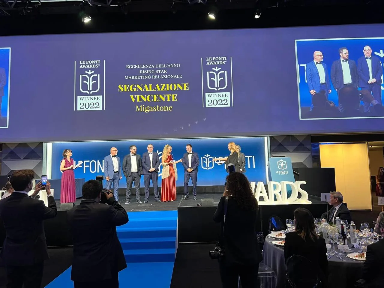 Il palco dei Le Fonti Awards 2022 durante la premiazione: sul maxischermo
                        la dicitura «Eccellenza dell'anno Rising Star Marketing Relazionale —
                        Segnalazione Vincente, Migastone», e il gruppo dei premiati sul palco