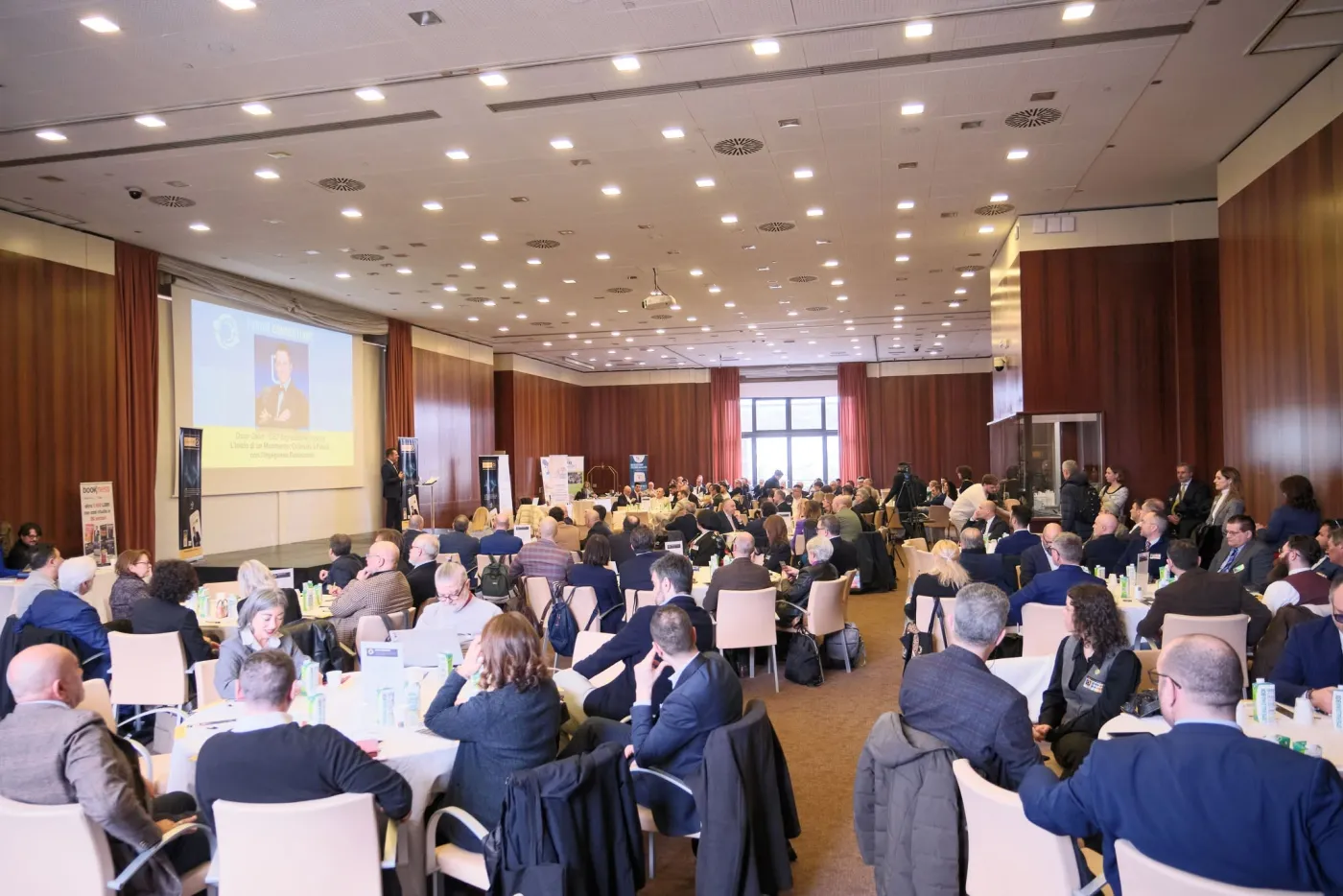 The Forum Connections hall in Florence during a plenary session: dozens of
                      entrepreneurs and professionals seated at round tables, with a speaker on
                      stage and a screen behind them