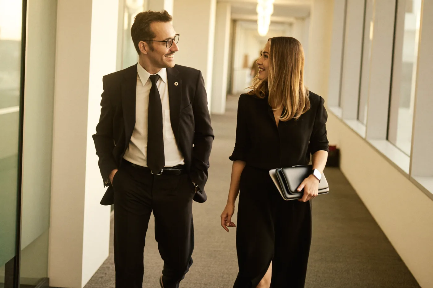 Two colleagues walking and smiling down an office
                      corridor while talking: the younger one on the left, a
                      senior on the right