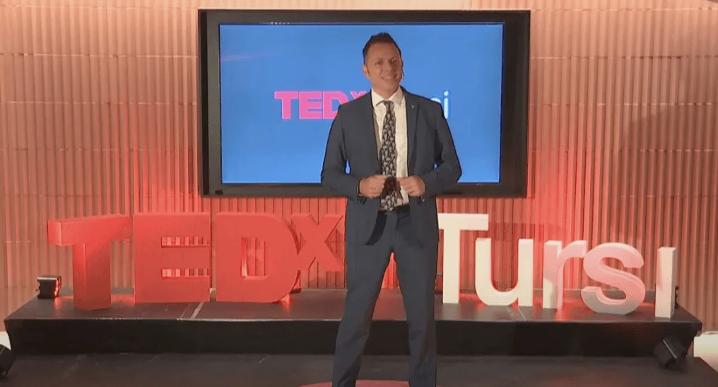 Oscar Dalvit sul palco di TEDx Tursi: in piedi davanti alle lettere
                        bianche e rosse dell'allestimento, con il proprio intervento proiettato
                        sullo schermo alle spalle