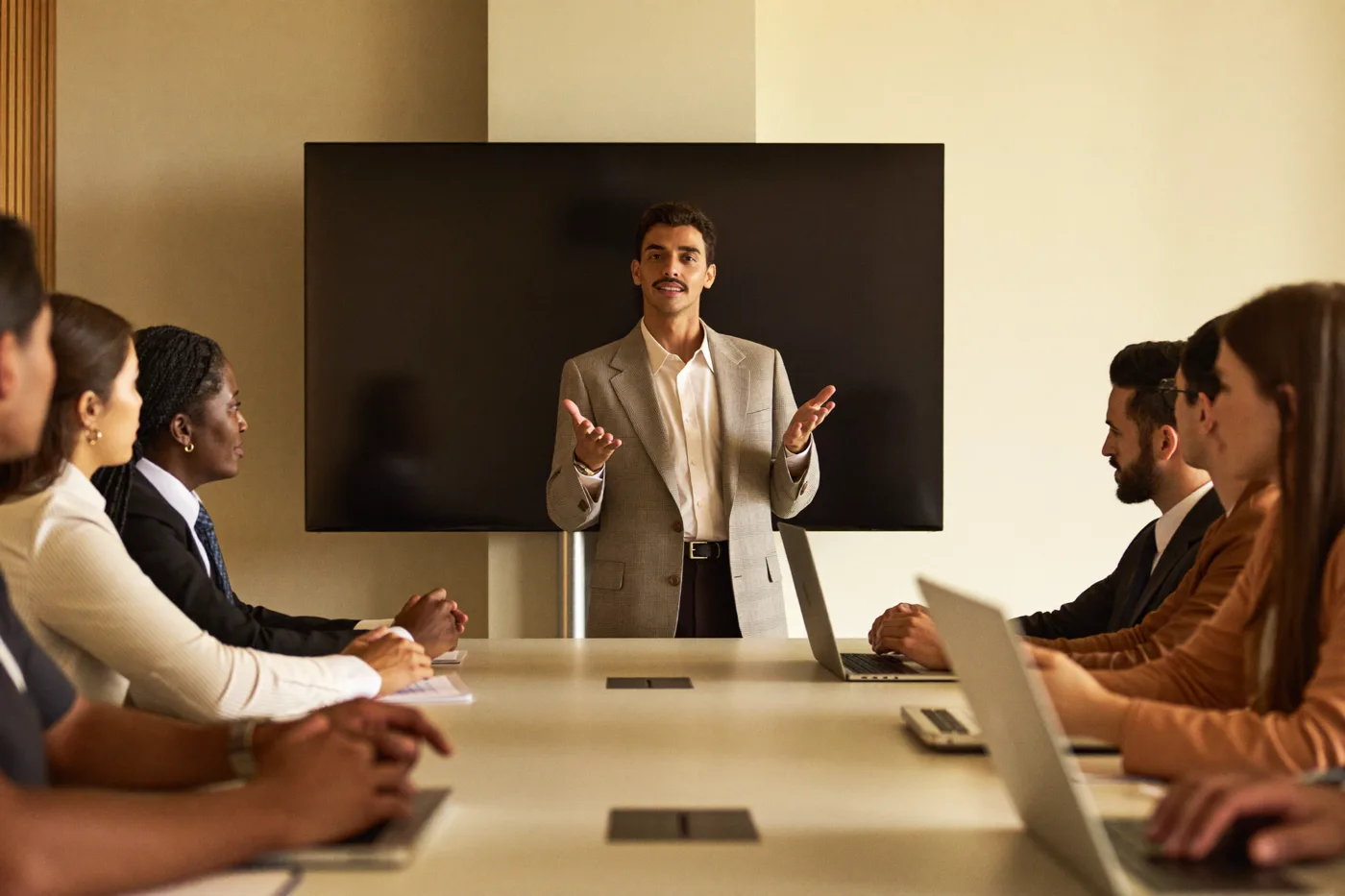 A coordinator standing in front of a table of agents with
                      laptops and notepads, presenting in a meeting room